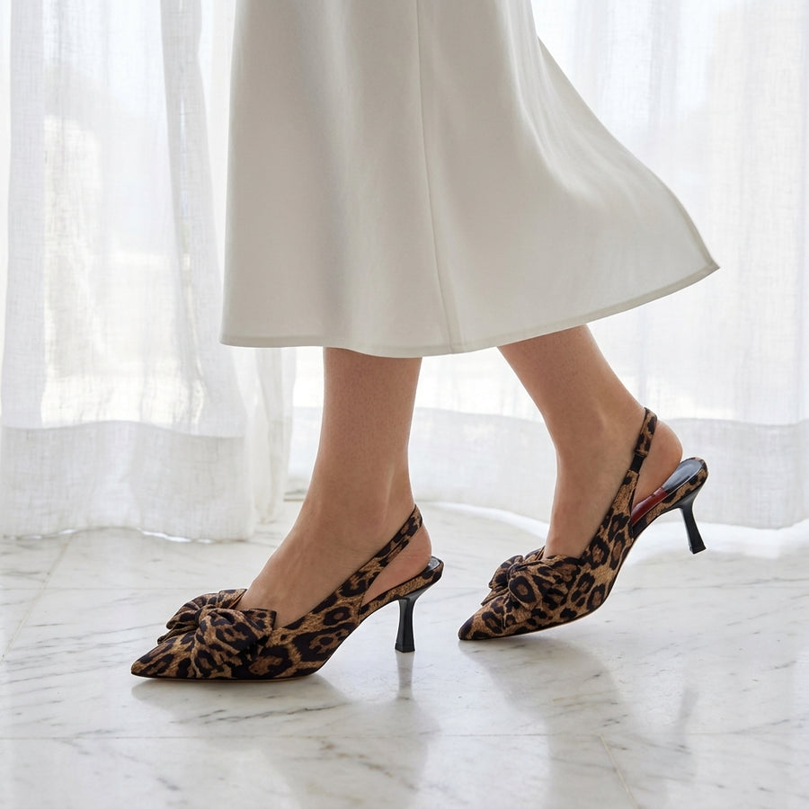 A pair of leopard-print pointed-toe slingback heels featuring a decorative bow on the front, worn with a white skirt on a marble floor.